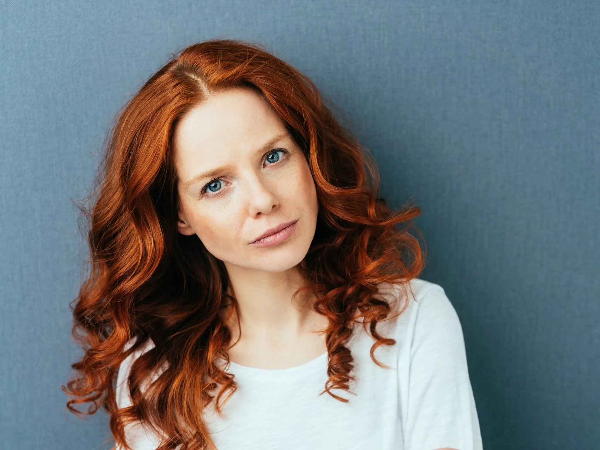 A woman with red hair looking thoughtfully at the camera against a blue wall